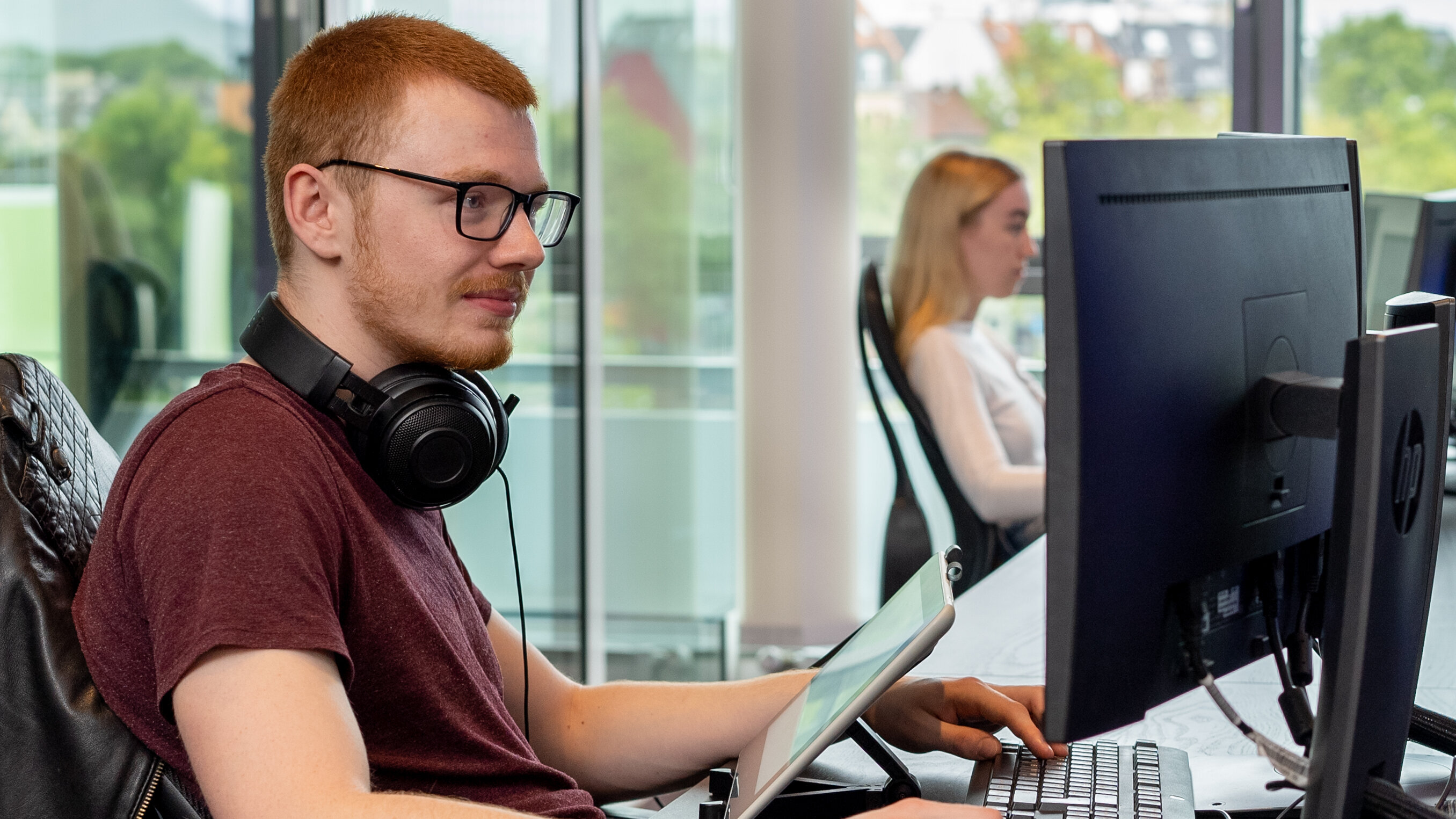 A young man wearing glasses and headphones works at a desk using a graphics tablet and a large monitor in a modern, light-filled room. Another person is working in the background.