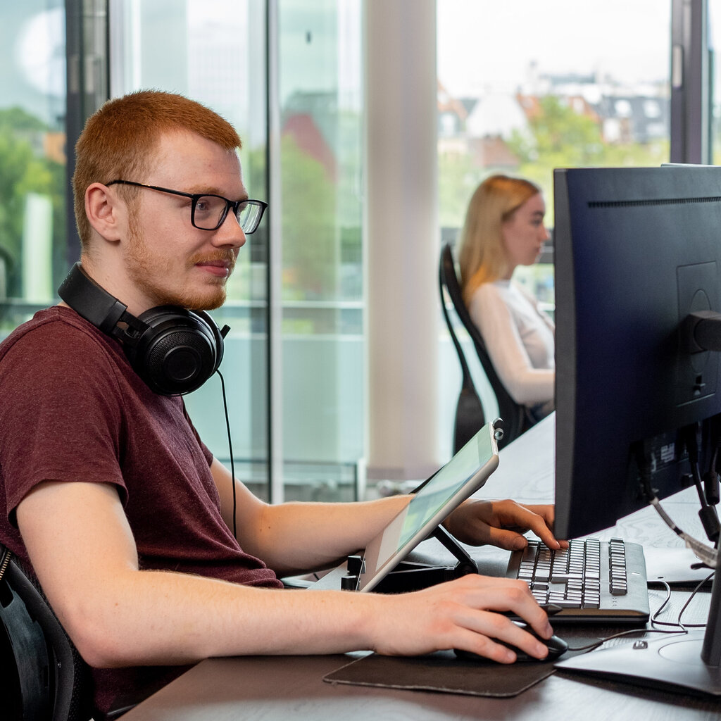A young man wearing glasses and headphones works at a desk using a graphics tablet and a large monitor in a modern, light-filled room. Another person is working in the background.