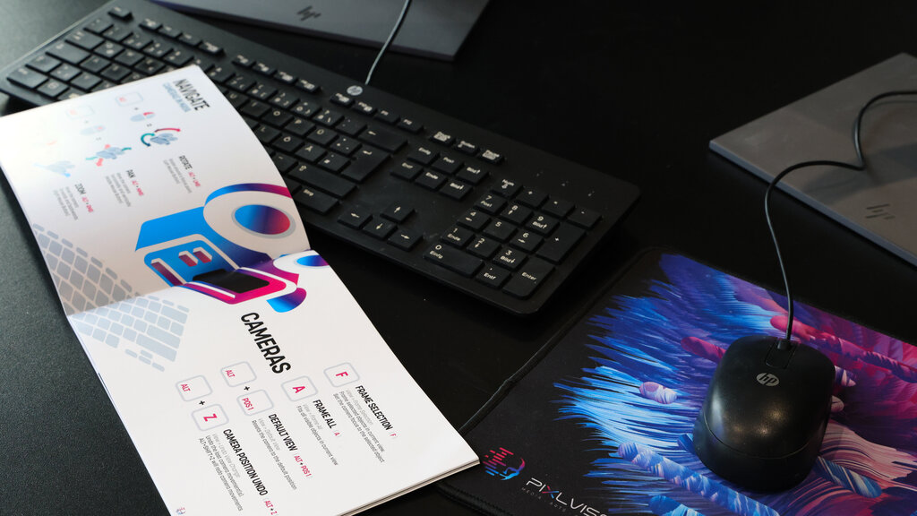 Close-up of a workspace featuring an HP keyboard, PIXL VISN mousepad, mouse, and an open booklet displaying colorful camera navigation tips.